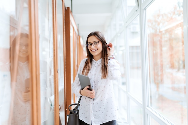 Portrait of collage girl standing next to noticeboard with tablet in arm and bag on the shoulder