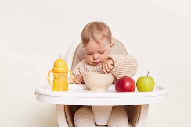 Portrait of concentrated baby wearing beige sweater sitting in a child's chair isolated on a white background holding spoon in hand and eating porridge with attentive expression