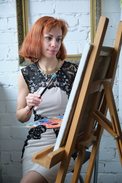 Portrait of concentrated beautiful woman painter holding art palette and painting on canvas with brush at home