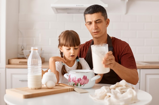 Portrait of concentrated dark haired man wearing maroon t shirt and apron cooking in kitchen with little daughter, holding capacity with milk, having serious expression.