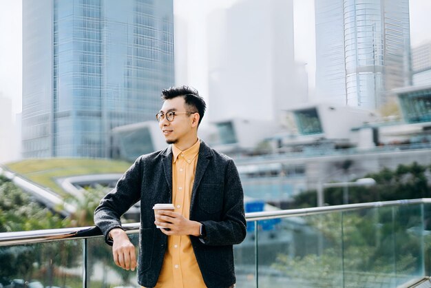 Photo portrait of confidence asian businessman looking away and having coffee to go against city scene in front of modern office buildings