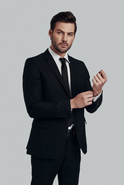 Portrait of confidence. Handsome young man in full suit adjusting sleeve and looking at camera while standing against grey background