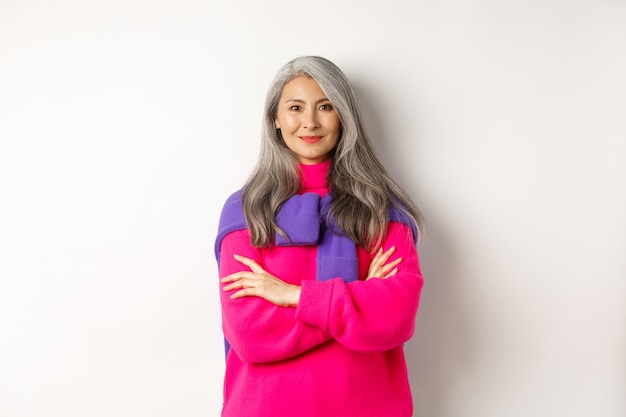 Portrait of confident asian senior woman cross arms on chest and smiling, standing over white background in trendy clothes