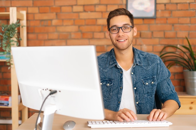 Portrait of confident cheerful handsome young freelance designer male in stylish glasses working on desktop computer sitting at desk at home office