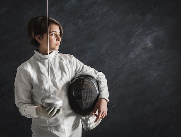 Photo portrait of confident fencer girl in white costume, holding protective mask and foil. black studio background, copy space