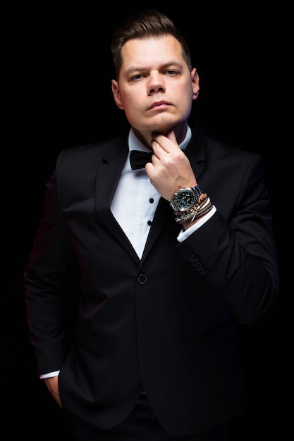 Portrait of confident handsome elegant stylish businessman with bow-tie holding hand near beard on black  in studio