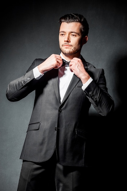 Portrait of confident handsome man in black suit with bowtie posing in dark studio background