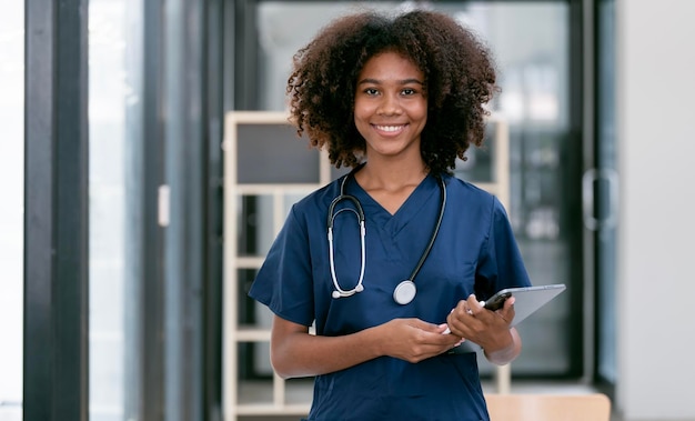 Portrait of confident happy female nurse or doctor with stethoscope and tablet standing in hospital hallway smiling to camera