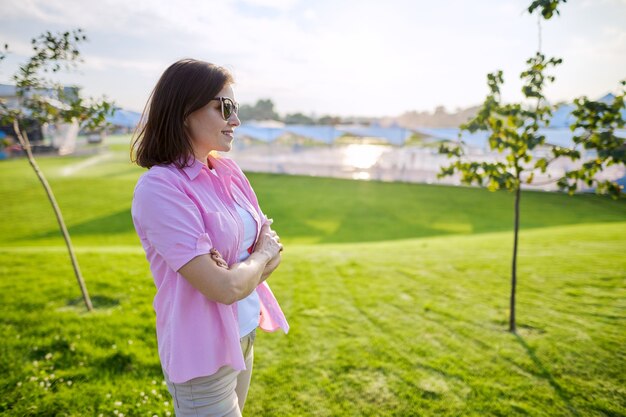 Portrait of confident mature woman with folded hands in shirt sunglasses, green grass lawn background copy space, sunset summer day