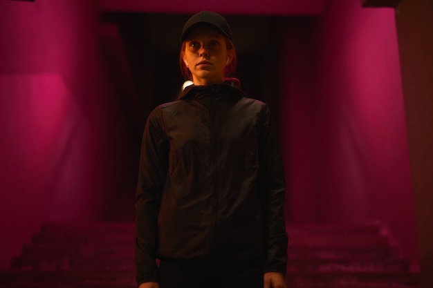 Portrait of confident redhead female in black cap and headphones walking outdoors in night city alon...