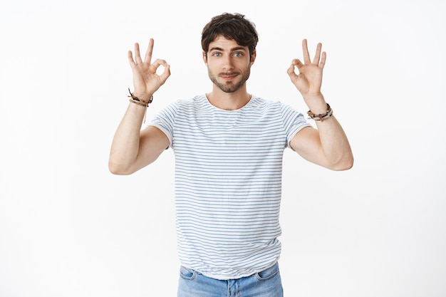 Portrait of confident and skillful good-looking male coworker with blue eyes and beard wearing striped t-shirt showing okay gestures with both hands assuring everything ok, project going perfect