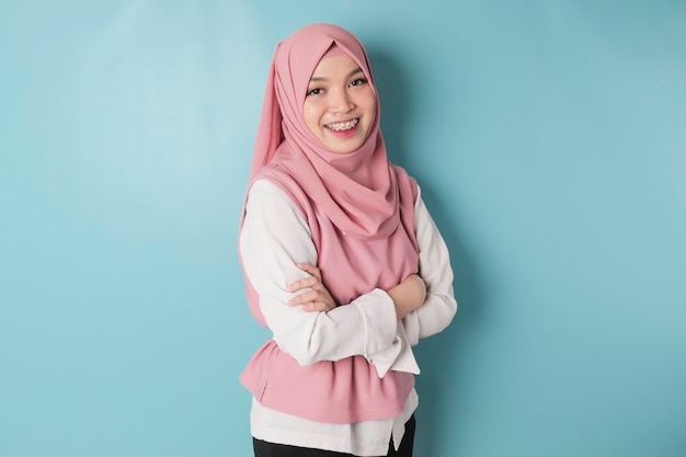 Portrait of a confident smiling Asian Muslim woman standing with arms folded and looking at the camera isolated over blue background