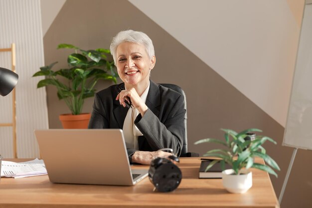 Portrait of confident stylish european middle aged senior woman at workplace Stylish older mature 60s gray haired lady businesswoman sitting in modern office Boss leader teacher professional worker