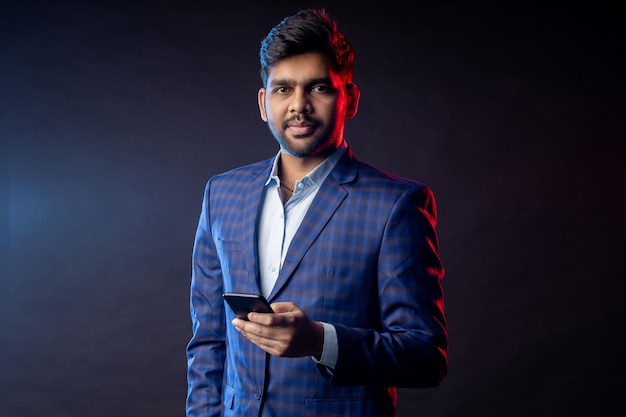 Portrait of confident stylish young indian man in formalwear, holding mobile phone, looking at screen, standing against black background. Successful businessman. Business, technology.