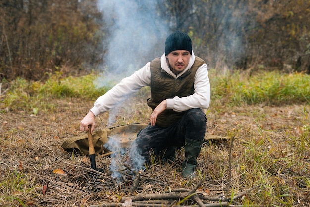 Portrait of confident tourist male wearing warm clothes sitting near burning campfire and holding small shovel, looking at camera. Concept of scout, research, travel and survival in nature.