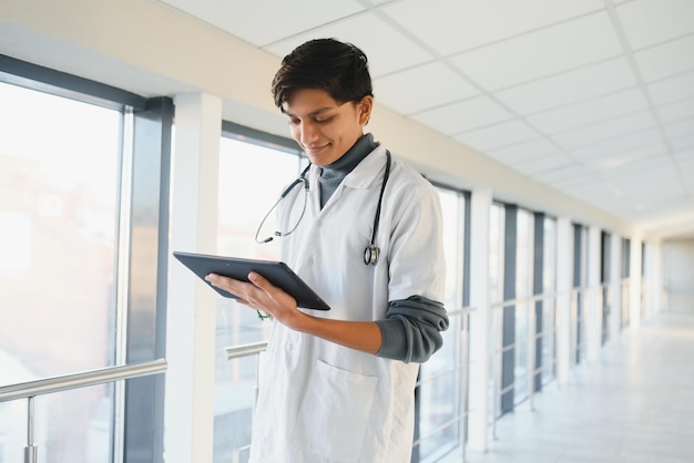 Portrait of confident young Arabian Indian male medical doctor in white coat, standing with clipboard in hands on background of modern hospital building outdoor