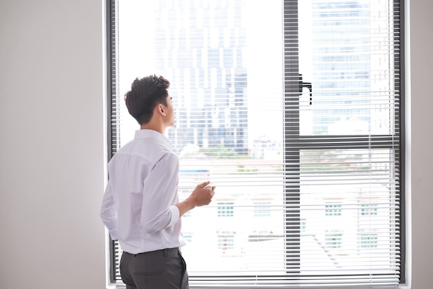 Portrait of a confident young businessman holding a cup of coffee while standing near office window, intelligent men in white shirt while resting after meeting