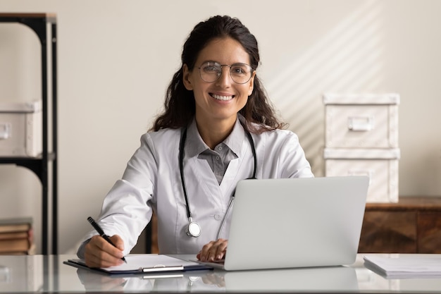 Portrait of confident young Caucasian female doctor in white medical uniform sit at desk working on computer Smiling woman GP or physician in glasses use laptop write in medical journal in clinic