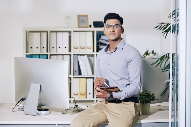 Portrait of confident young Indian entrepreneur sitting on edge of office desk with tablet computer in hands and looking at camera