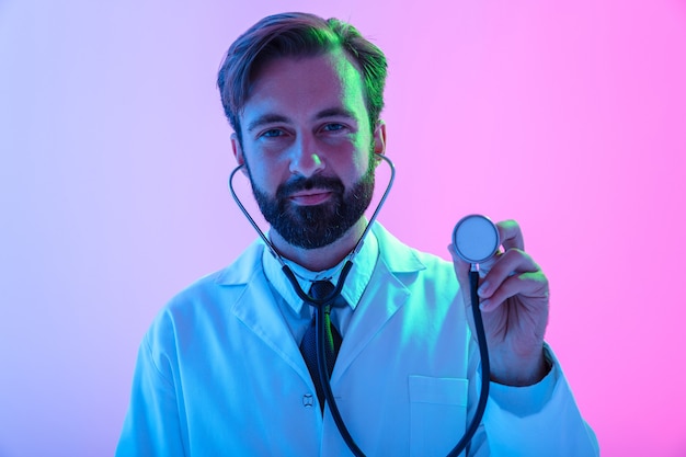 Portrait of a confident young man doctor wearing uniform standing isolated over pink-blue haze , using stethoscope