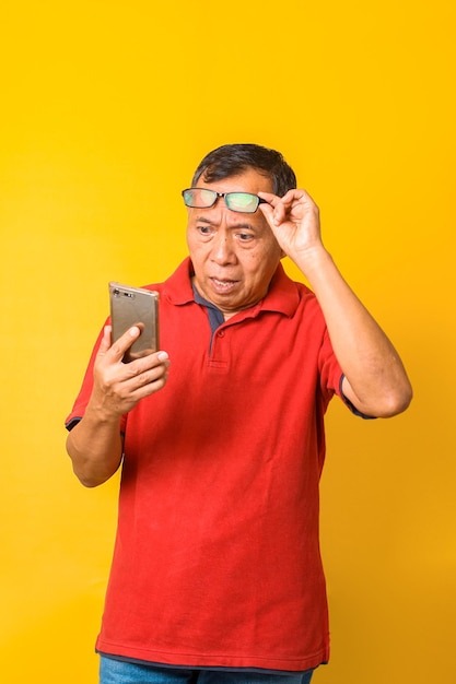 Portrait of confused Asian old man looking on smartphone while putting on glasses isolated on yellow