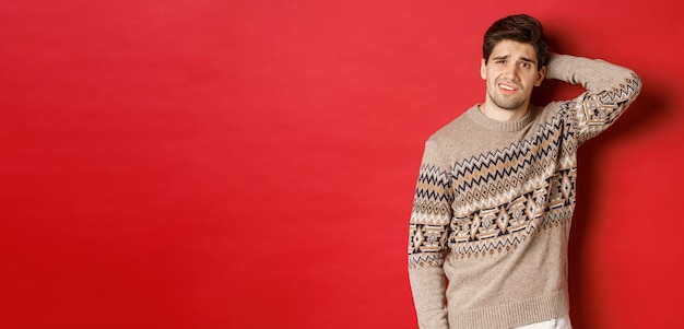 Portrait of confused man in christmas sweater scratching back of head and looking clueless about new year presents standing over red background