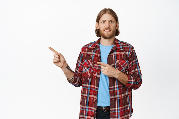 Portrait of confused nervous blond man, pointing fingers left and frowning, cringe in doubt disbelief, standing in casual outfit against white background.