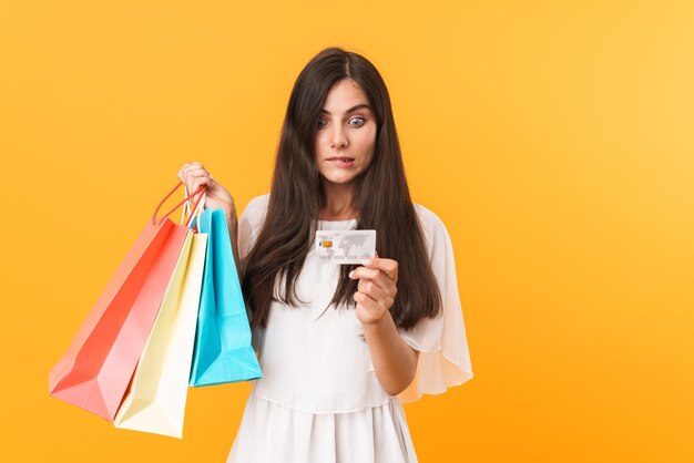 Portrait of confused shopaholic woman wearing dress holding credit card and colorful shopping bags isolated over yellow wall