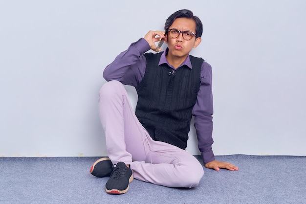 Portrait of Confused young Asian man talking on mobile phone while sitting on floor with legs crossed isolated over white background