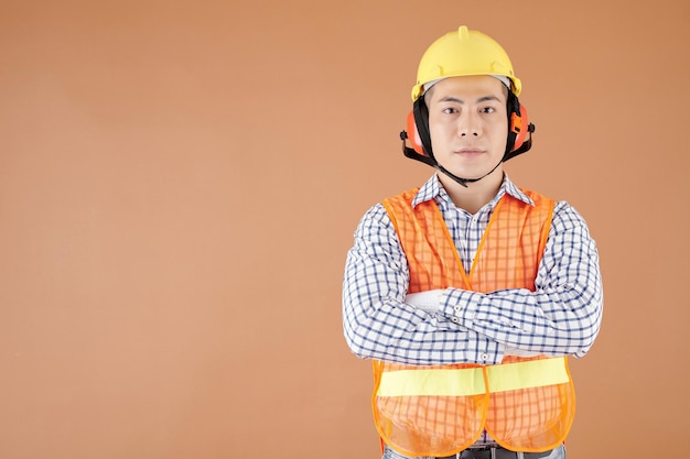 Portrait of construction worker in bright vest hardhat and earmuffs crossing arms and looking at camera