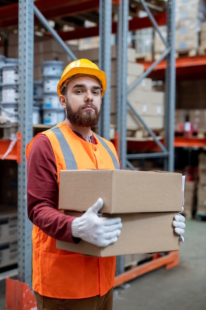 Portrait of content confident young depository mover in hardhat and reflective vest standing with boxes against warehouse frame