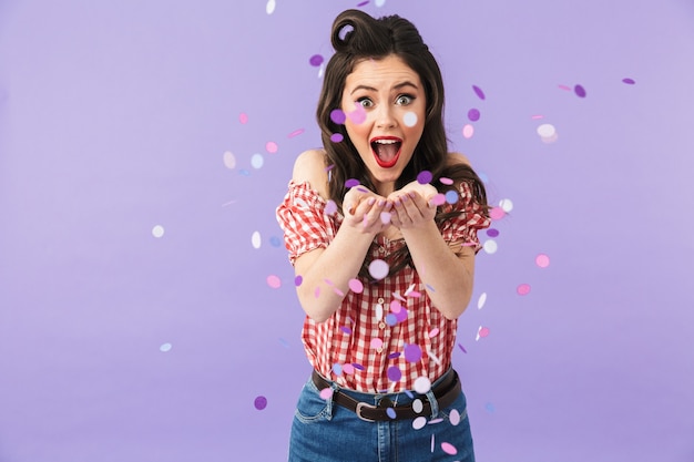 Portrait of content pin-up woman in american style wear smiling and celebrating holiday under falling confetti isolated over violet wall