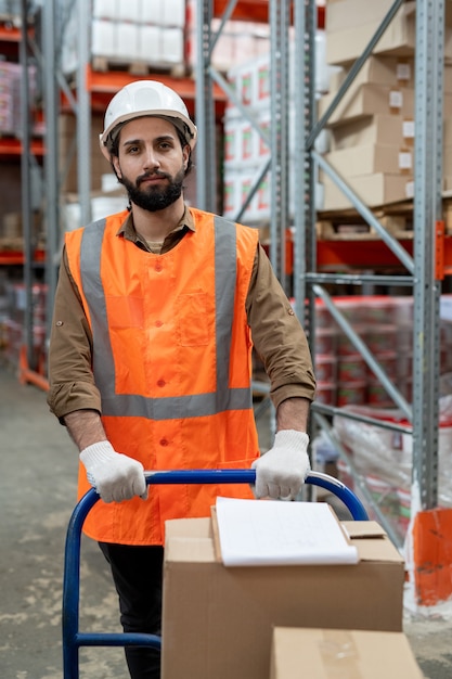 Portrait of content young mixed race man in reflective vest and hardhat pushing storage cart over warehouse