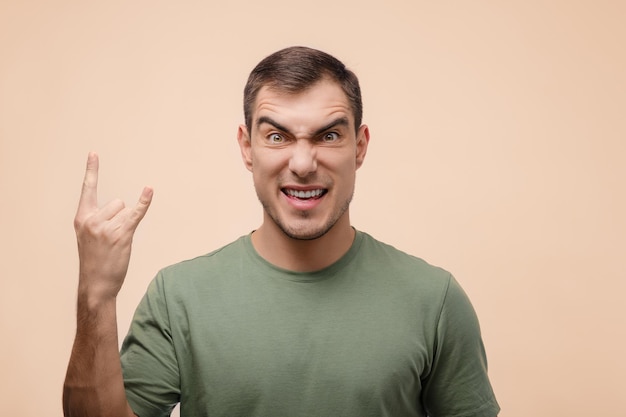 Portrait of contented man with short hair shows goat gesture horns on beige background fullface