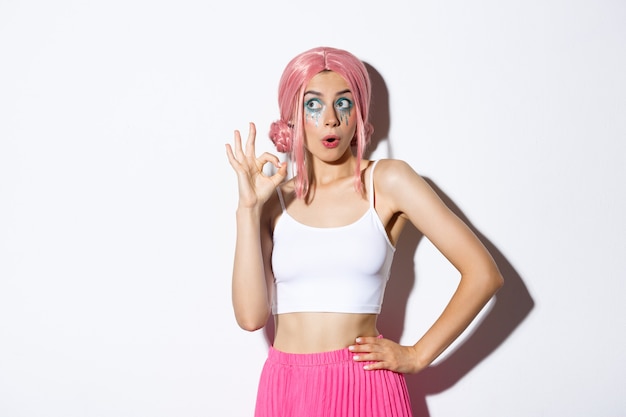 Portrait of coquettish party girl in pink wig and halloween outfit, showing okay sign and looking at upper left corner advertisement, standing over white background