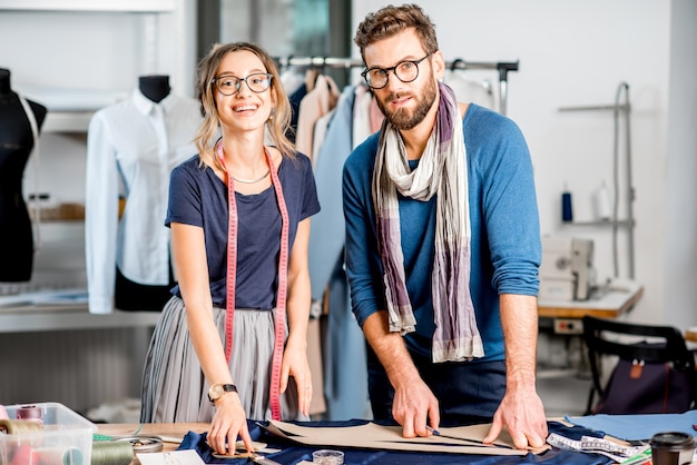 Portrait of a couple of fashion designers working with fabric and clothing sketches at the studio full of tailoring tools and equipment