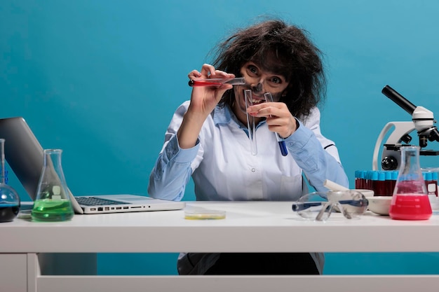 Portrait of crazy chemist with wild look mixing dangerous chemical compounds while sitting at desk on blue background. Mad woman scientist with funny face expression creating new toxic formula.