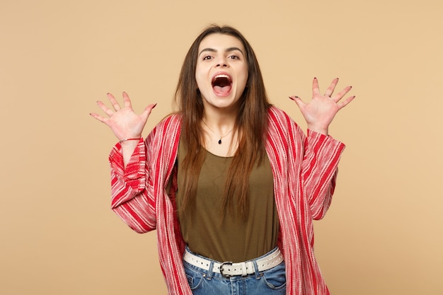 Portrait of crazy young woman in casual clothes spreading hands, keeping mouth wide open isolated on pastel beige background in studio. People sincere emotions, lifestyle concept. Mock up copy space.