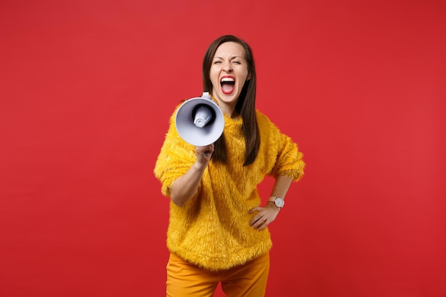 Portrait of crazy young woman in yellow fur sweater standing, screaming on megaphone isolated on bright red wall background in studio. People sincere emotions, lifestyle concept. Mock up copy space.