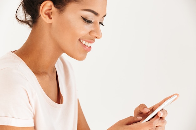 Portrait cropped of mixed race woman in casual typing or using smartphone holding in hands, isolated over white wall