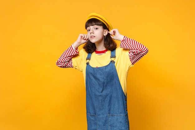 Portrait of curious girl teenager in french beret, denim sundress looking attentively far away distance isolated on yellow background. People sincere emotions, lifestyle concept. Mock up copy space.