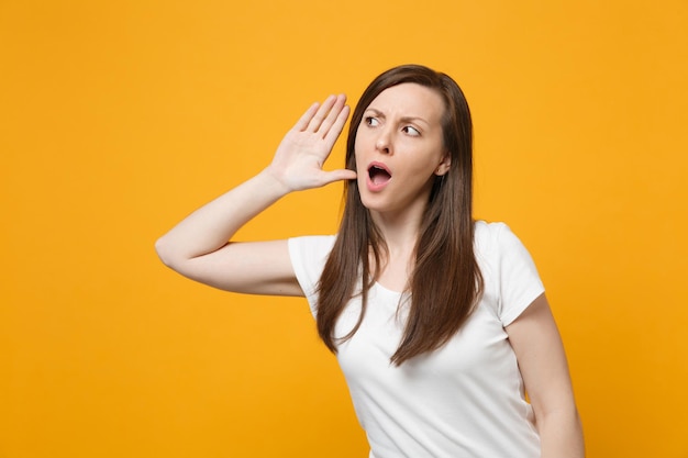 Portrait of curious young woman in white casual clothes looking aside try to hear you with hand near ear isolated on yellow orange background in studio. People lifestyle concept. Mock up copy space.