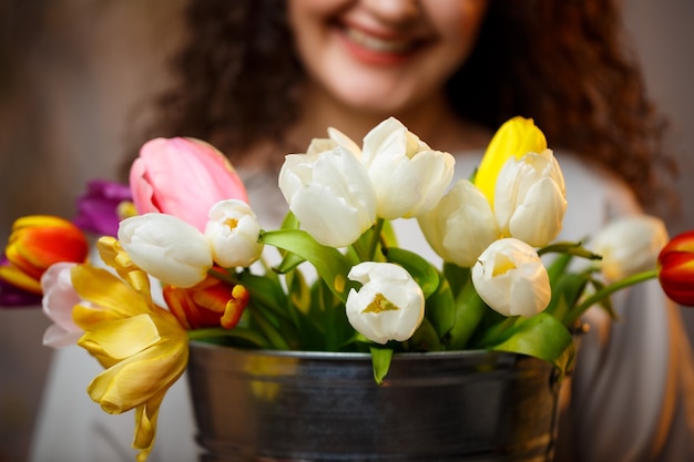 Portrait of a curly girl with a bucket of tulips. Fresh flowers for beautiful photos. Delicate photos with flowers in the studio