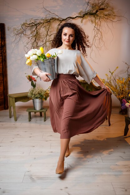 Portrait of a curly girl with a bucket of tulips. Holds fresh flowers and goes for beautiful photos. Bright photos with flowers in the studio