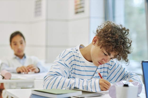 Portrait of curly hair young boy sitting at desk in school classroom and writing copy space