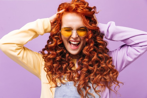 Portrait of curly naughty redhead girl in yellow sunglasses posing on isolated wall