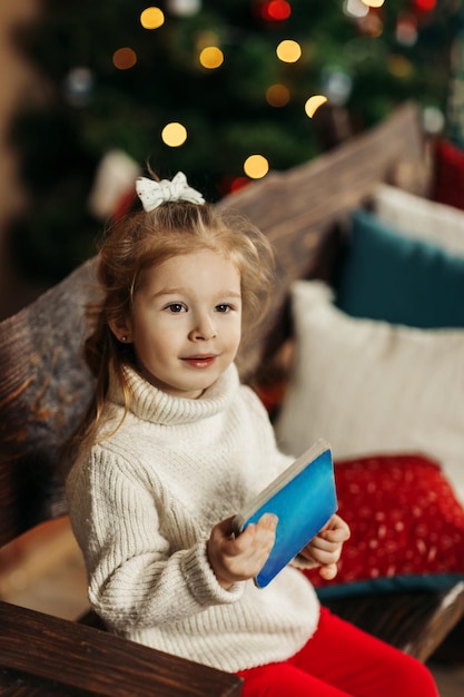 Portrait of a curlyhaired little girl in a warm knitted sweater with a book in her hands against the background of a Christmas tree enjoying a cozy winter evening at home Christmas New Year