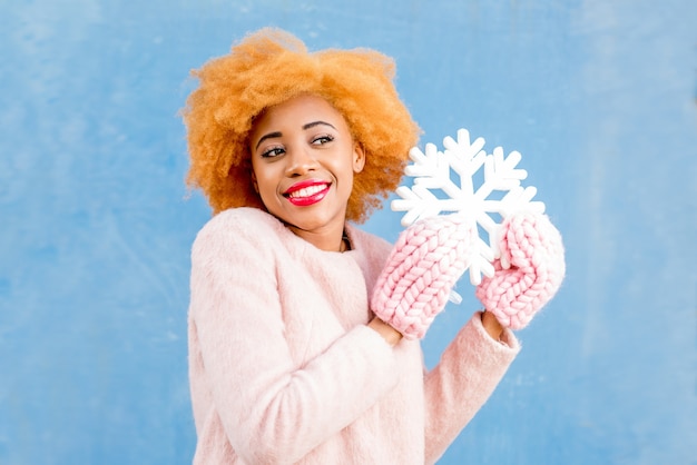 Portrait of a cute african woman in the pink coat holding an artificial snowflake on the blue wall background. Winter season concept