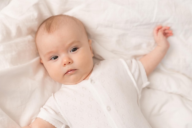 portrait of a cute baby in a white bodysuit on a bed at home with white linens. Newborn baby at home