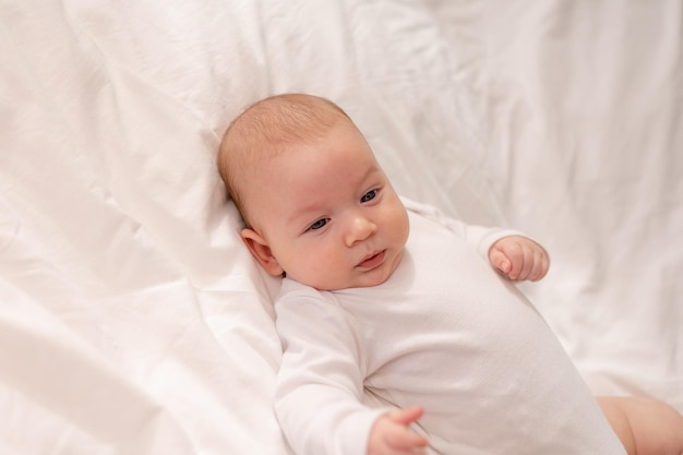 Portrait of a cute baby with blue eyes in a white bodysuit lying on the back on white bed linen